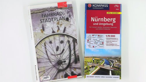 Zwei Fahrrad-Stadtpläne für Nürnberg liegen nebeneinander auf weißem Grund: links ein Plan des ADFC mit einem künstlerischen Fahrradspeichen-Motiv und rechts eine KOMPASS-Fahrradkarte für Nürnberg und Umgebung.